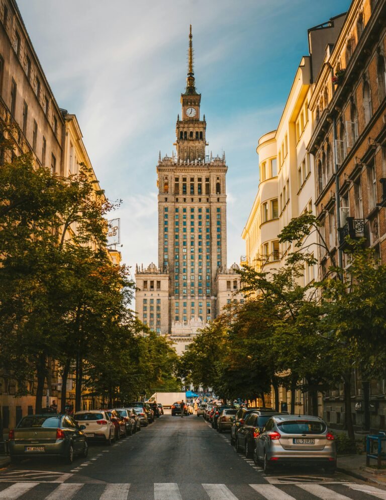 Zdjęcie Pałacu Kultury w oddali na tle prawie bezchmurnego nieba. Na pierwszym planie ulica w perspekrywie zbieżnej po lewej i prawej stronie drzewa.